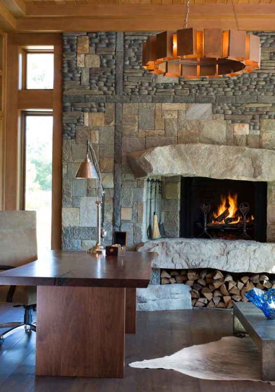 Rustic-modern stone fireplace with a thick limestone mantel, glowing fire, and adjacent wood desk in a warm, modern home office