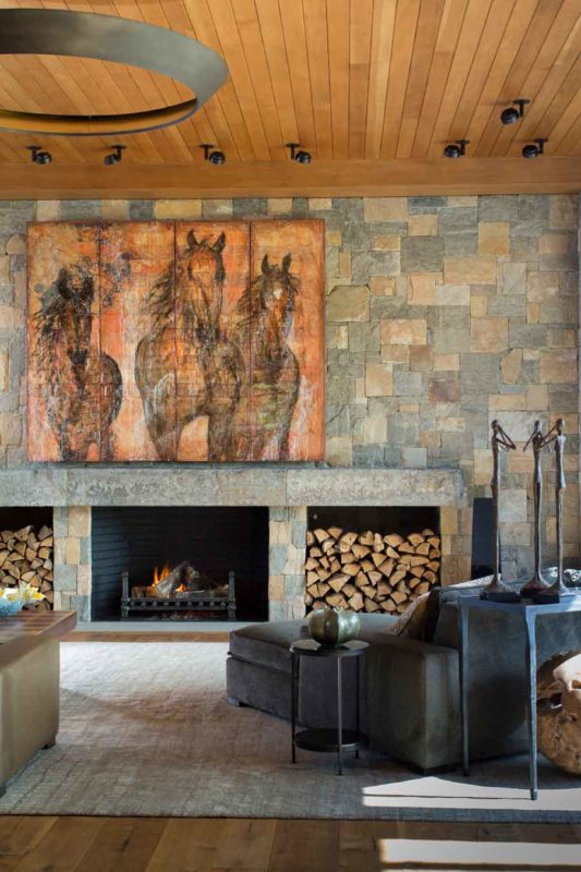 Contemporary living room featuring a wide stone fireplace, stacked firewood niches, and large-scale horse artwork beneath a wood-paneled ceiling