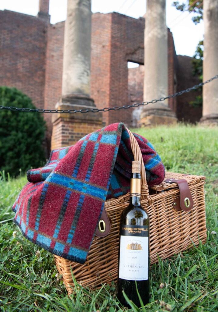 A bottle of Vermentino Reserve wine by Barboursville Vineyards and a picnic hamper and picnic blanket ready for a winery picnic near the Barboursville ruins.