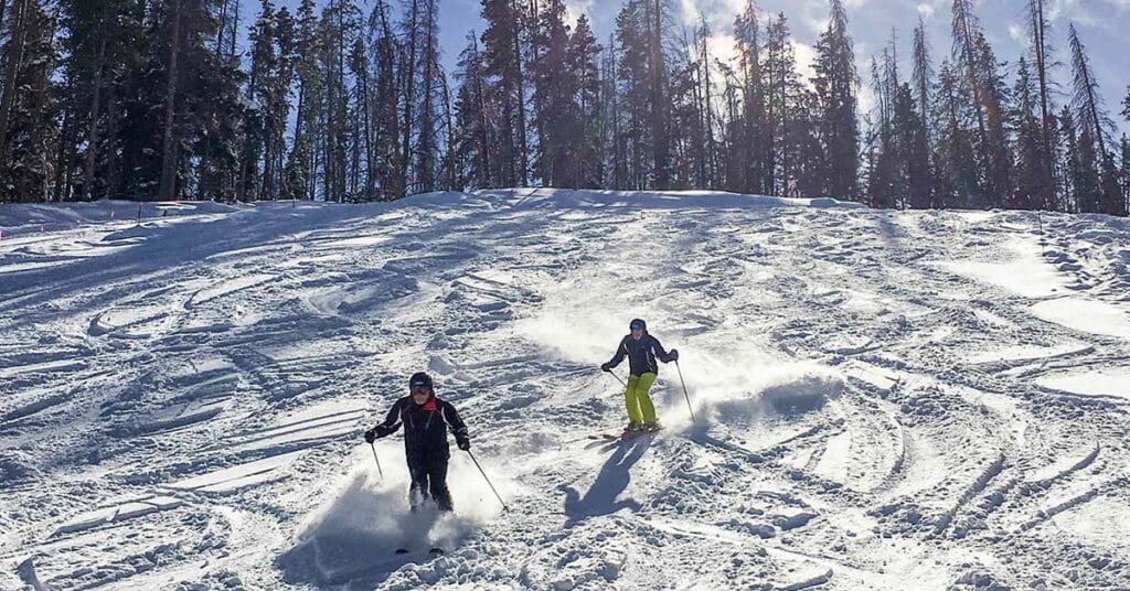 Skiers descending a powder-covered slope at Vail Resort, Colorado, with the sun reflecting on the mountains.