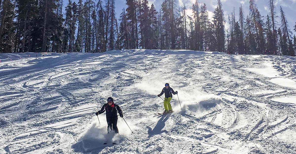 Skiers descending a powder-covered slope at Vail Resort, Colorado, with the sun reflecting on the mountains.