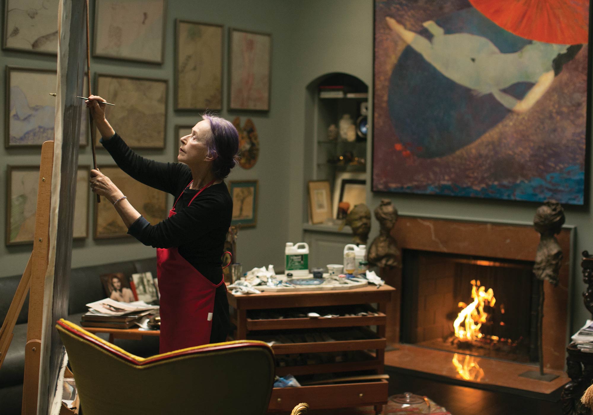 Beatrix Ost painting in her art studio in Charlottesville with paint lining the walls and a fireplace in the background
